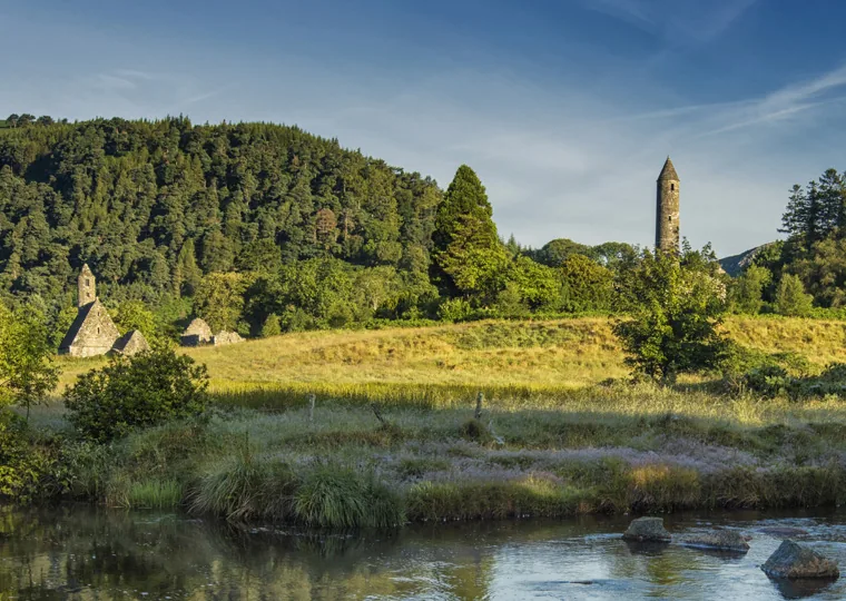 Glendalough St Kevin's Kitchen and round tower reflected beside a calm river, forested hills rising behind